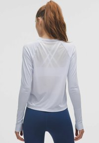 Hosszú ujjú fehér sportpóló, könnyű anyagból készült, átlátszó hátsó panellel, keresztbe fonott mintával és hüvelykujjlyukas mandzsettákkal.