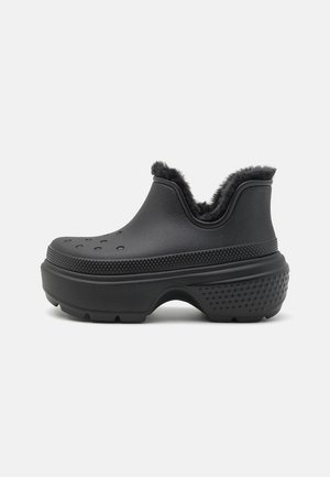 Scarpa nera senza lacci con suola di gomma testurizzata, bordi rialzati, fori di ventilazione e una morbida fodera in pelliccia isolante all'apertura.