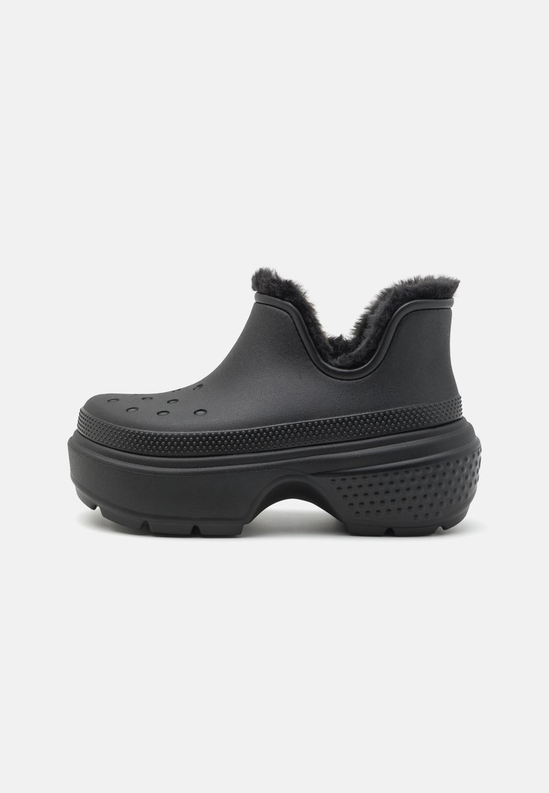 Shoe preto tipo slip-on com sola de borracha texturizada, bordas elevadas, orifícios de ventilação e um forro de pelo acolchoado e isolante na abertura.