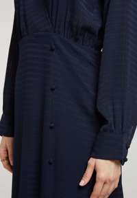 Robe bleu marine en tissu texturé, avec une taille froncée, des manches longues et de petits boutons recouverts de tissu sur le côté.