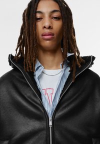 Svart jacka med dragkedja och foder av päls, lager på lager över en blå- och vitrandig skjorta och en vit t-shirt med tryck. Silverkedja synlig.