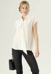 Una camicia bianca a maniche corte realizzata in un tessuto morbido e strutturato; presenta un dettaglio frontale drappeggiato. Abbinata a jeans neri aderenti e a una piccola borsa nera.