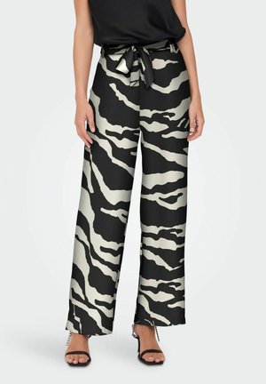 Vrouw met een broek met hoge taille en wijde pijpen met zebraprint en een gestrikte tailleband, zwart mouwloos topje en zwarte open-toe hakken met enkelkettingen.