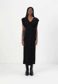 Black, floor-length dress with a V-neckline and cap sleeves. Smooth fabric with a slight sheen. Worn with black, knee-high boots.