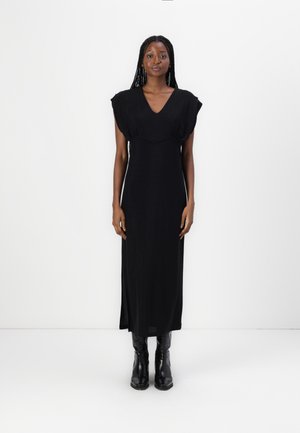 Black, floor-length dress with a V-neckline and cap sleeves. Smooth fabric with a slight sheen. Worn with black, knee-high boots.
