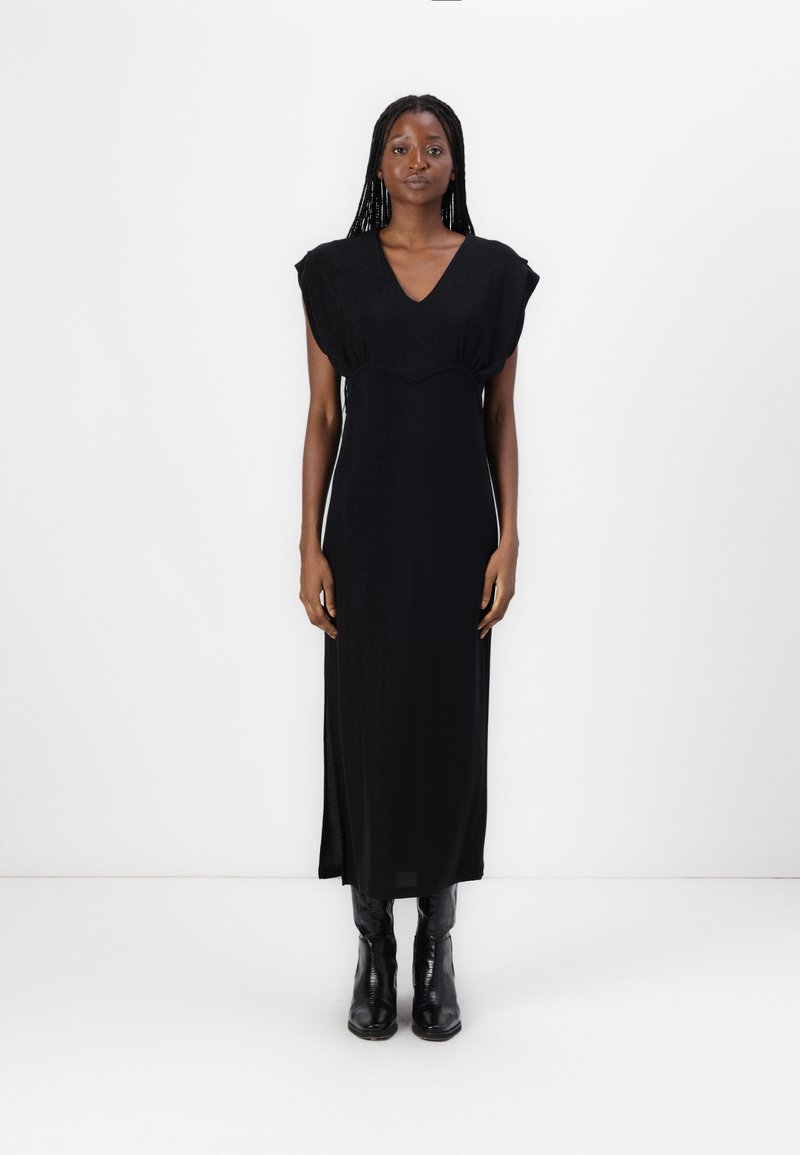 Black, floor-length dress with a V-neckline and cap sleeves. Smooth fabric with a slight sheen. Worn with black, knee-high boots.