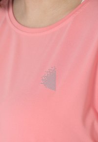 Camiseta deportiva de color rosa claro fabricada con una tela suave, que presenta un sutil gráfico gris en forma de corazón, ubicado en el área del pecho.
