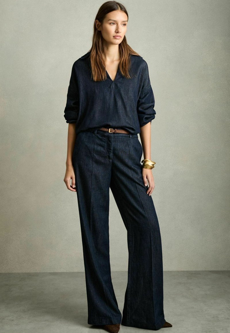 Dunkles Denim-Outfit mit einem locker sitzenden, langärmeligen Hemd und weitem Hosenbein. Enthält einen braunen Gürtel und ein goldenes Armreif.