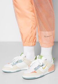 Sneakers med vita, krämfärgade och pastellfärgade detaljer; mesh-texturerade persikofärgade byxor; elastiska muddar; diskret Nike-logotyp på byxorna och skorna.