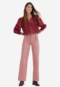 Blouse rouge texturée avec des manches bouffantes et des boutons, associée à un jean large taille haute de couleur rose pâle. Modèle portant des bottines noires.