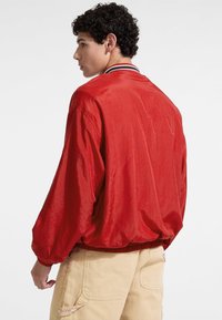 Giacca bomber rossa con una texture lucida, caratterizzata da maniche lunghe, colletto a coste e orlo elastico. Abbinata a pantaloni beige chiaro.