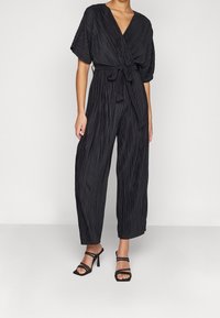 Combinaison noire plissée avec un décolleté en V profond, des manches larges, une taille à nouer et des jambes larges, associée à des talons noirs à lanières.