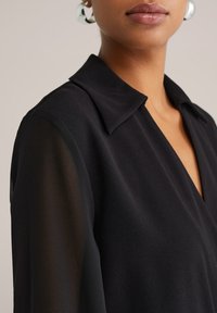 Čierna blúzka s golierom a hlbokým výstrihom do V, priesvitnou látkou na rukávoch a mäkkým, textúrovaným materiálom.