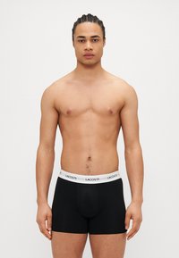 Fekete boxer alsónadrág fehér elasztikus derékpánttal, amelyen a "LACOSTE" logo található. Puha anyag, testhezálló forma és elülső zsebe a támogatás érdekében.