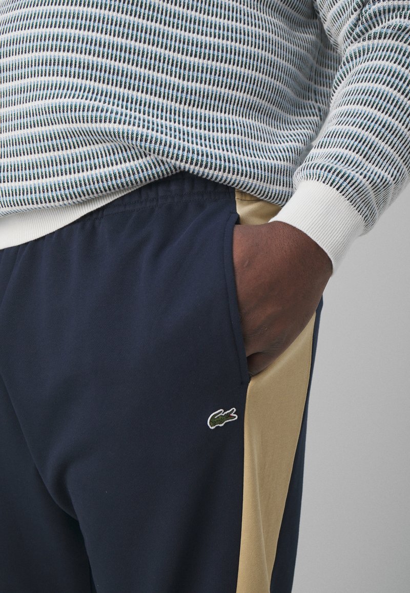 Pantalon de jogging bleu marine avec un panneau latéral beige et un logo crocodile vert brodé. Poignets côtelés blancs sur un pull léger à motifs au-dessus.