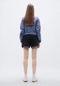 Γαλάζιο κοντό μπουφάν από ελαφρύ ύφασμα, σε συνδυασμό με μαύρο σορτς. Το μοντέλο φοράει ανοιχτόχρωμα παπούτσια με κάλτσες, εμφανίζεται από πίσω με φόντο μία απλή επιφάνεια.
