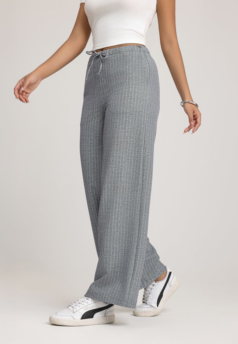 Grijs gestreepte witte pijpenbroek gemaakt van lichte stof, met een elastische tailleband en witte sneakers als schoeisel.