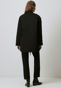 Cappotto nero con colletto alto, maniche lunghe e vestibilità rilassata. Abbinato a pantaloni neri a gamba dritta e stivaletti.