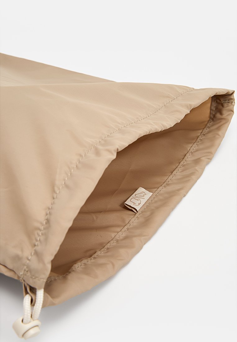 Manga de chaqueta beige con puño elástico y cordón ajustable que presenta un tope de plástico sobre un fondo blanco.