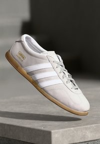 Sapatilha de camurça cinza claro com design de três riscas brancas, biqueira redonda, sola de borracha e o logótipo "Gazelle" em dourado na lateral.