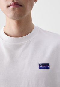 T-shirt branca de algodão com decote redondo e mangas curtas. Apresenta um patch retangular azul e vermelho no peito.