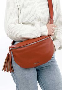 Borsa a tracolla in pelle marrone con finitura texturizzata, chiusura con zip e dettaglio a frange. Indossata sopra un maglione bianco e jeans.