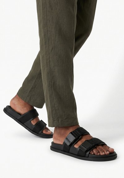 Pieds portant des sandales noires à bouts ouverts avec deux sangles réglables et un pantalon vert foncé sur un fond blanc uni.
