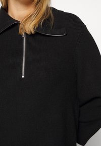 Svart ribbad tröja med hög krage, halvmatchande dragkedja framtill med silvriga detaljer och lösa ärmar. Mjuk textur.