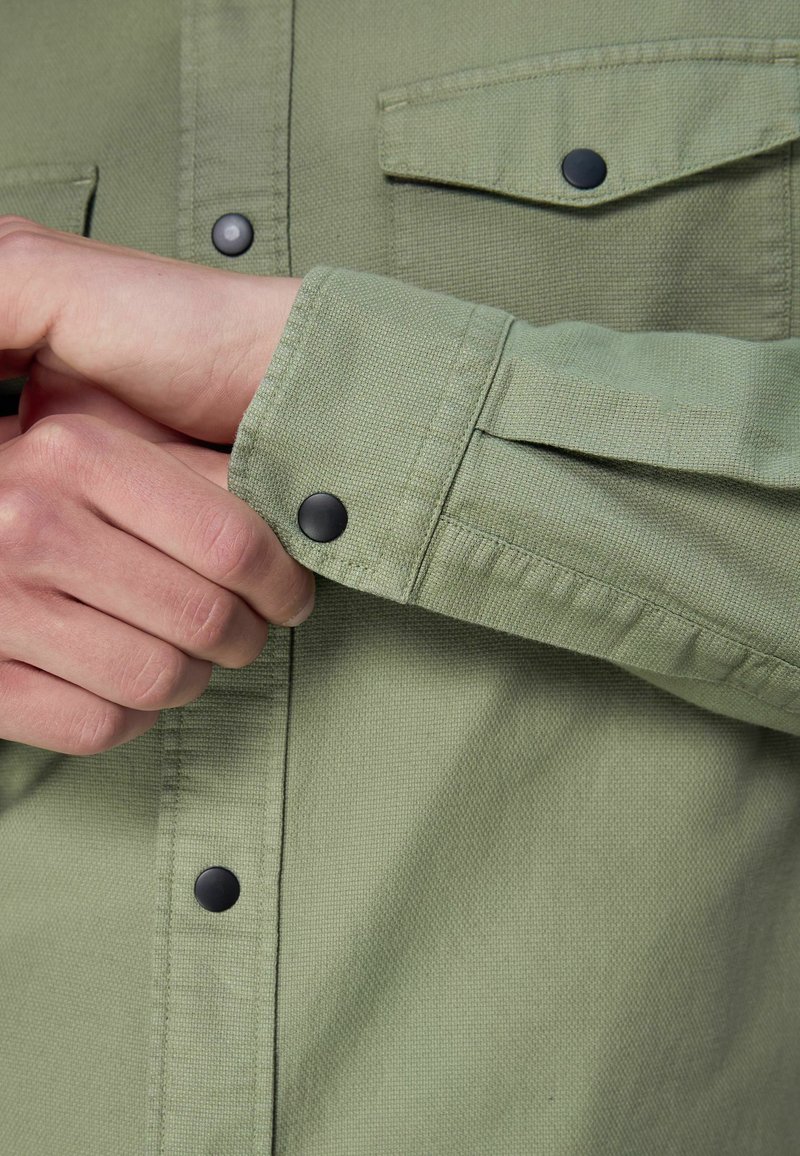 Green woven shirt featuring a textured finish, black snap buttons, and two chest pockets with flap closures. Cuff detail visible.