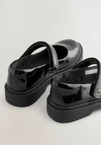 Pantofi din piele la denumire neagră, cu vârf rotunjit, curea la gleznă și talpă groasă din cauciuc negru. Prezintă un accent în formă de stea pe curea.