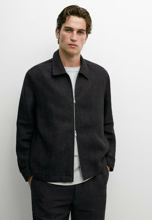 Jeune homme aux cheveux courts et ondulés, portant une veste noire zippée sur une chemise blanche, debout devant un fond blanc uni.