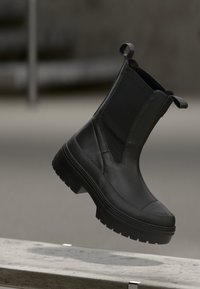 Cizme negre până la gleznă, cu o talpă plată și groasă, un sistem superior din piele netedă, panouri laterale elastice și tab-uri pentru o îmbrăcare mai ușoară.