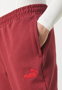 Bordeauxfarbene Jogginghose aus strukturiertem Stoff, mit elastischem Bund, seitlichen Taschen und einem roten gestickten Logo am linken Oberschenkel.