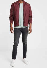 Burgundowa kurtka bomber, jasnoniebieska koszulka, ciemne dżinsy, białe trampki. Kurtka ma ściągacze i zamek błyskawiczny. Prosty, nowoczesny krój.