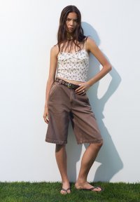 Brune denim bermuda shorts med en afslappet pasform, parret med en tætsiddende hvid top med blomsterprint og et mørkt bælte med cirkulært hardware.
