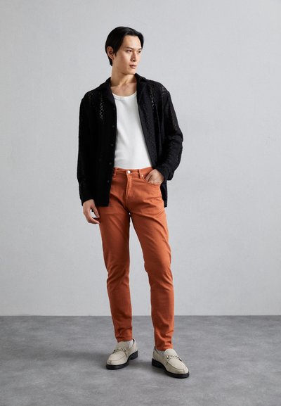 Svart spets-knappskjorta, vit T-shirt med rund hals, orange slim fit-byxor och ljusgrå loafers med metallaccent.