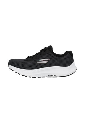 Zapatilla deportiva negra con parte superior de malla, suela blanca acolchada y detalle de logo. Presenta un diseño con cordones y patrón texturizado en el lado.