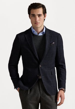 Mørk marineblå rutejakke med slank pasform, to-knap lukning og notch-lapel. Kombineret med en grå sweater og en lyseblå skjorte med krave.