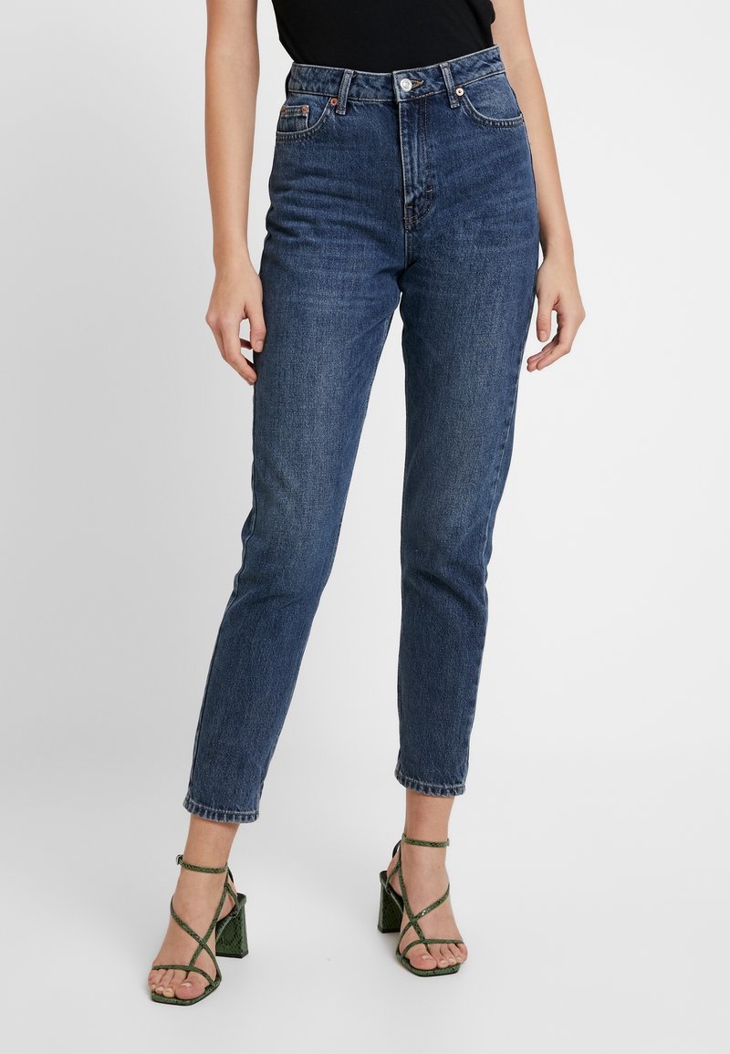 Personne portant un jean slim bleu foncé taille haute et des sandales vertes à talons hauts avec des brides croisées sur fond blanc.