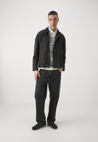 Črna lahko jakna z žepi spredaj, nošena preko črtastega polo majice, v kombinaciji s temnimi jeans hlačami in črnimi supergami. Nevtralna ozadje.