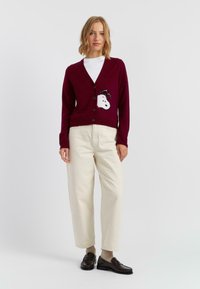 Bordeaux cardigan met een witte cartoonhond met een zwarte sjaal op de voorkant, gecombineerd met crèmekleurige wijde broek en donkere instappers.
