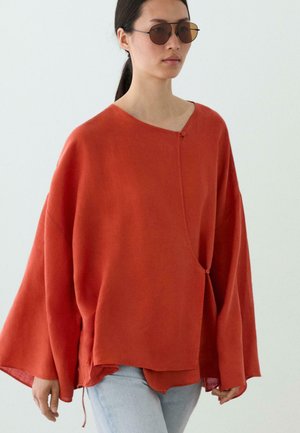 Jeune femme portant un haut orange rouille oversize avec une fermeture asymétrique à boutons et un jean bleu clair, portant également des lunettes de soleil teintées marron.
