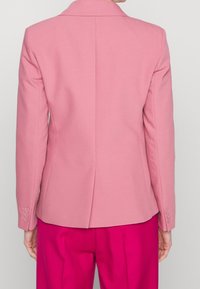 Blazer rosa a medida hecho de tejido texturizado, con solapa tipo notch, ventilación posterior y acentos de cuatro botones en las mangas.