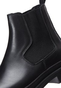 Botte de cheville en cuir noir avec panneaux latéraux élastiques, surface lisse et détails de couture texturée. Semelle robuste avec une légère élévation.