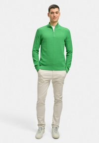 Grön stickad tröja med kvartslång dragkedja, ribbade muddar och nederkant, kombinerad med beige slim fit-byxor och ljusa sneakers mot en enfärgad bakgrund.