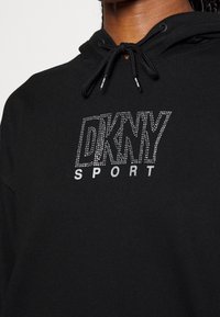 Svart hoodie i mjukt tyg, med dragsko i huvan och en silverfärgad rhinestone-logotyp "DKNY SPORT" på framsidan.