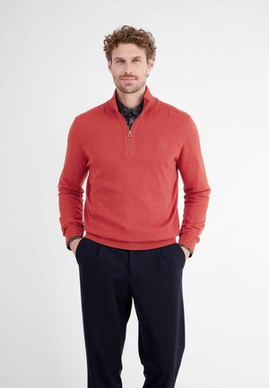 Man with curly hair wearing a red quarter-zip sweater over a dark shirt and black pants, standing with hands in pockets against a white background.