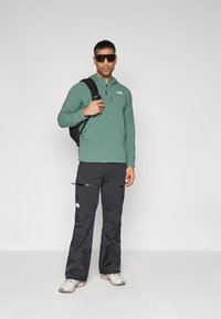 Chaqueta verde con cremallera y acentos negros, combinada con pantalones negros con bolsillos con cremallera. Llevada con una mochila negra y zapatos grises.
