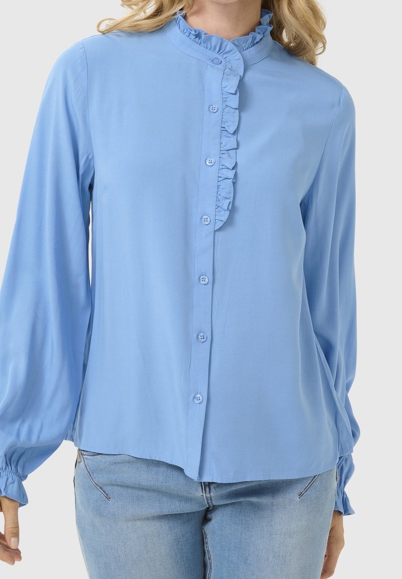 Femme portant un chemisier bleu clair à manches longues avec un col haut volanté et une patte de boutonnage à l'avant, associé à un jean bleu clair.