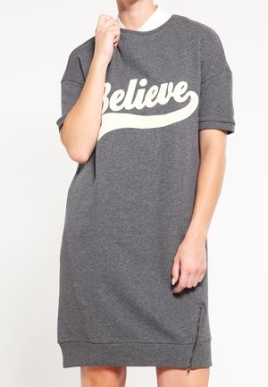 Kobieta ubrana w szarą sukienkę z krótkim rękawem, z napisem „Believe” w białym kolorze na przodzie oraz z małym zamkiem bocznym przy brzegu sukienki.